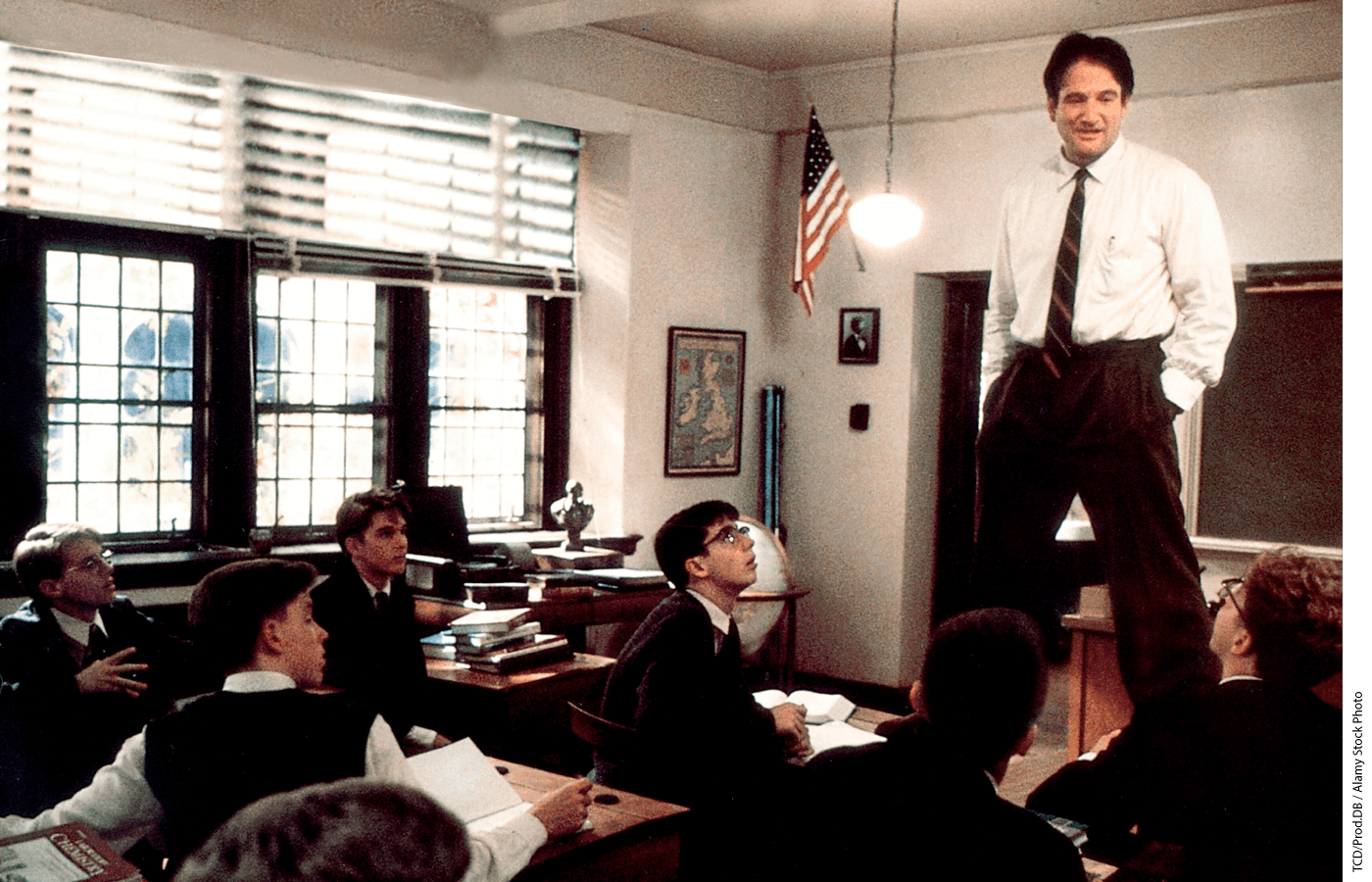 A Still from Dead Poets Society. Robin Williams as Mr. Keating straddles students' desks while delivering another big ol' dollop of inspiration